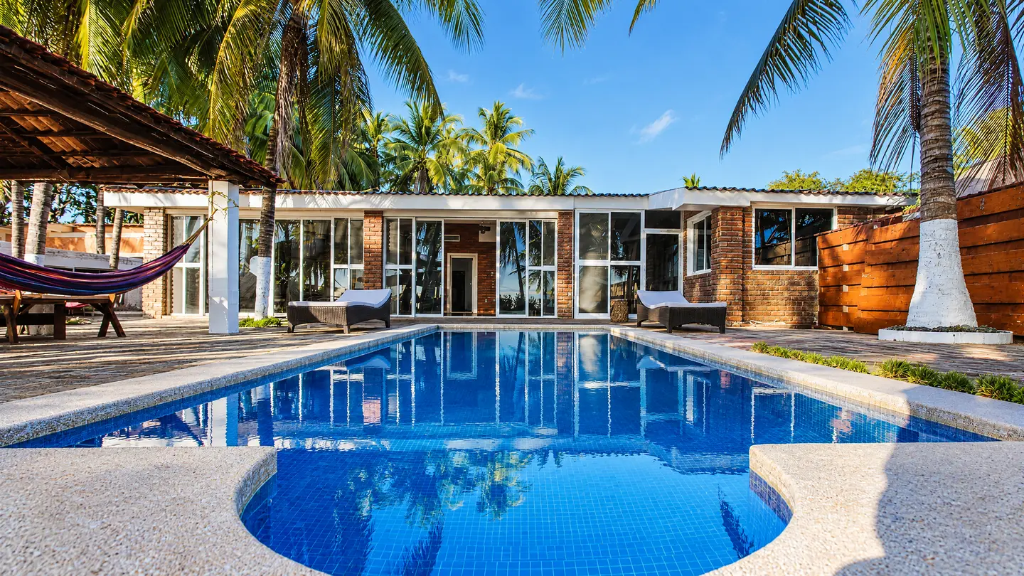Piscina azul frente a la casa, entre palmeras y cielo despejado